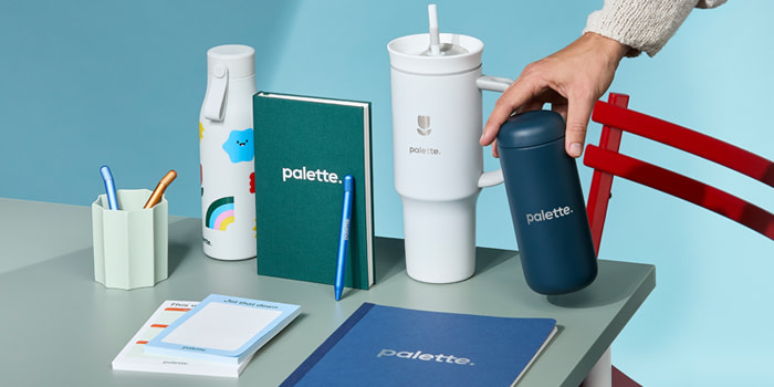 A variety of branded merch on a light blue surface, including a Hardcover Notebook, Tumblers, Twist Pen, Notepad and Water Bottle.