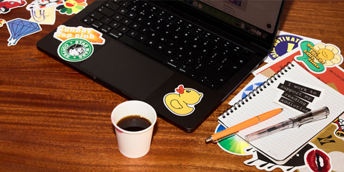 A collection of Die-cut Stickers on a brown desk, a black laptop and a paper cup filled with coffee.