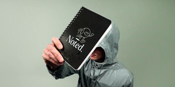 A person wearing a green rain jacket, holding up a black Spiral Notebook.