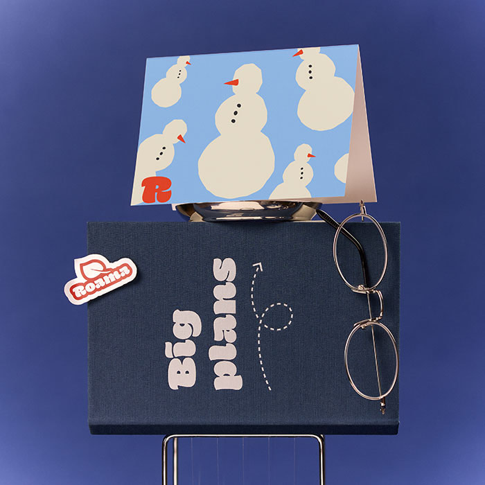 A branded holiday card on display on top of a branded Hardcover Notebook, with a branded Sticker and silver spectacles.