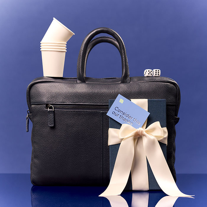 A black laptop bag with a Hardcover Nptebook in front of it, a Thank You Card and paper cups