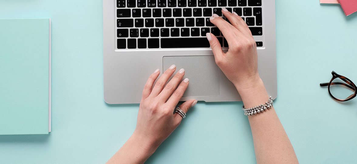 Flat lay picture of blue desk with notebook and glasses as well as hands typing on laptop
