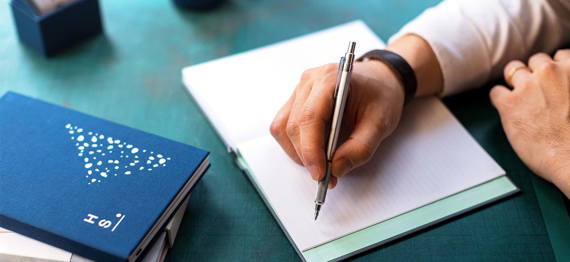 Hand writing on a MOO Hardcover Notebook at a green desk with another custom cloth cover notebook