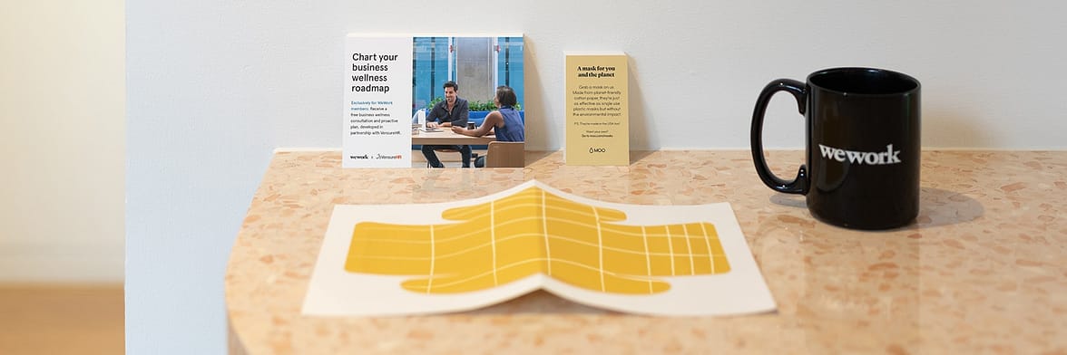WeWork yellow paper face mask on a desk with a postcard and a business card by MOO