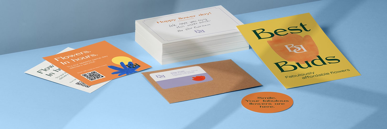 Orange and yellow flyers, postcards and round sticker next to a pile of white postcards and a brown craft envelope by fictional brand Best Buds