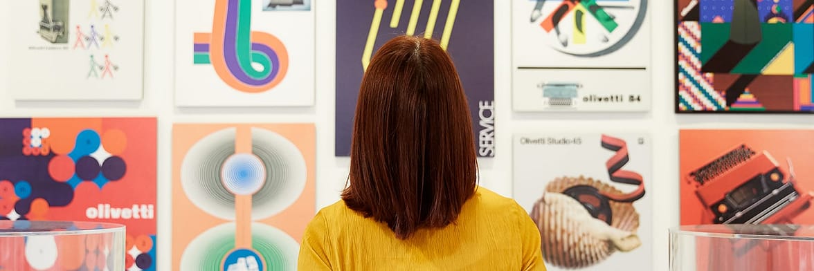 Red-haired woman seen from the back looking at a wall covered in design posters