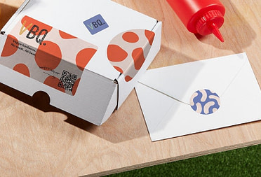 Box covered in colorful stickers, envelope with round sticker, sheet of mini stickers, ketchup, spatula, and barbecue food on a table