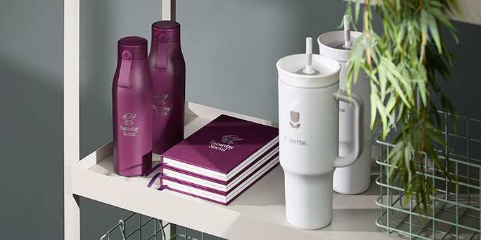 Branded purple Water Bottles, alongside purple Hard cover Notebooks and two white Tumblers on display.