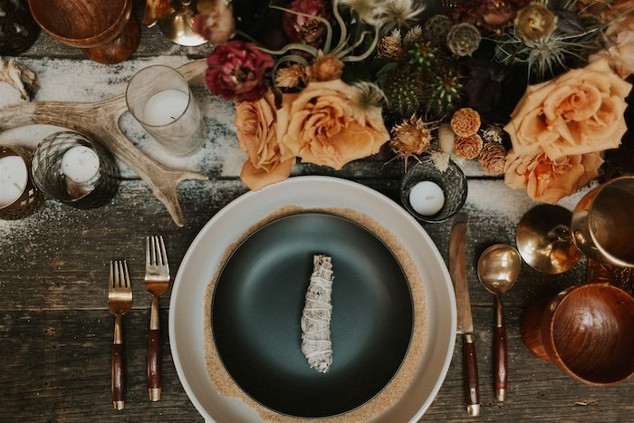 Tied dried leaves in a black plate surrounded by copper cutlery on a wooden table with candles, deer antlers and floral arrangements by The Floral Craft