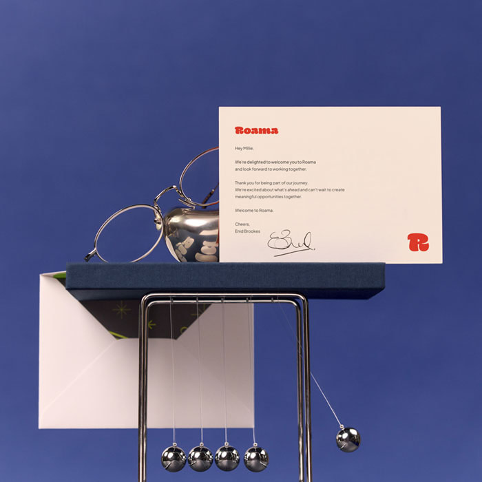 A branded cream-colored Thank You card displayed on a dark blue Hardcover Notebook, a silver decorative item, spectacles, with an opened envelope in the background. Everything is stacked on a silver newtons cradle.
