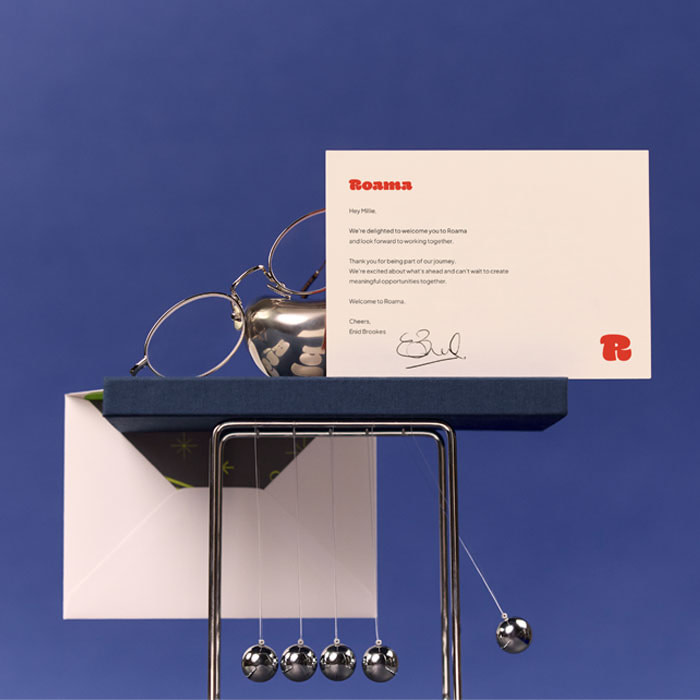 A branded cream-colored Thank You card displayed on a dark blue Hardcover Notebook, a silver decorative item, spectacles, with an opened envelope in the background. Everything is stacked on a silver newtons cradle.