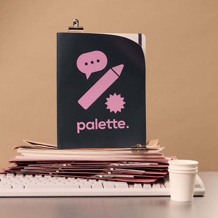 A branded pink and black Notebook, displayed upright on a stack of paper and a keyboard, with some empty paper cups displayed on the right side.