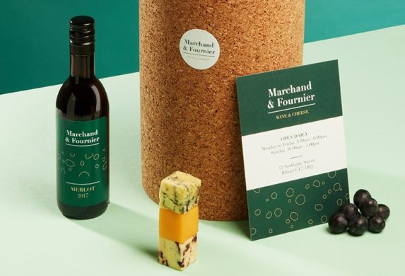 Mini bottle of wine with a green label, block of cheese, big cork pedestal with a round sticker, grape and big green postcard for a wine and cheese business