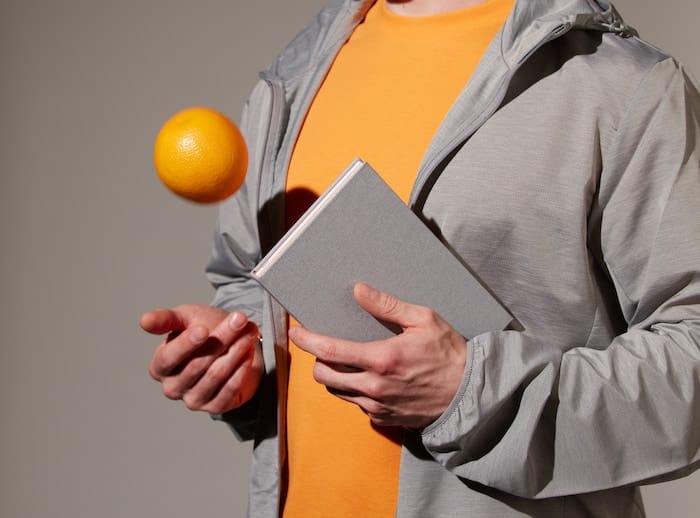 Person dressed in grey and orange holding a grey notebook and juggling with an orange