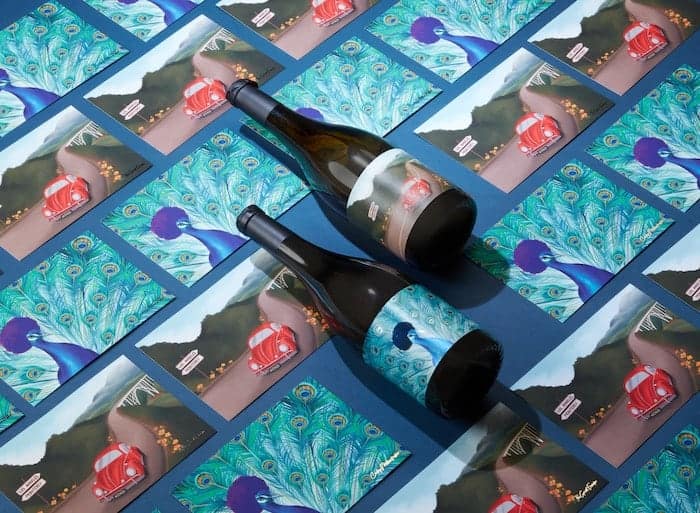 Two bottles of McBride Sisters Reserve Collection wine on a mosaic of MOO postcards with illustrations mirroring the wine labels. The first illustration is depicting a peacock with an afro and the other is a red beetle on the road
