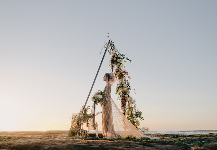 Woman in cream-colored wedding dress with a flower bouquet at dawn, standing in the middle of a triangle formed by a triangle canopy with flowers all over one side. The sky is clear and she's looking straight in front of her.