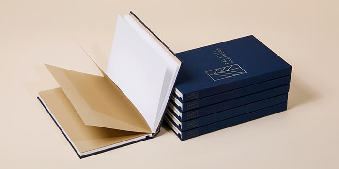 Open notebook next to a pile of five dark blue custom hardcover notebooks with a branded gold foil logo on the cover
