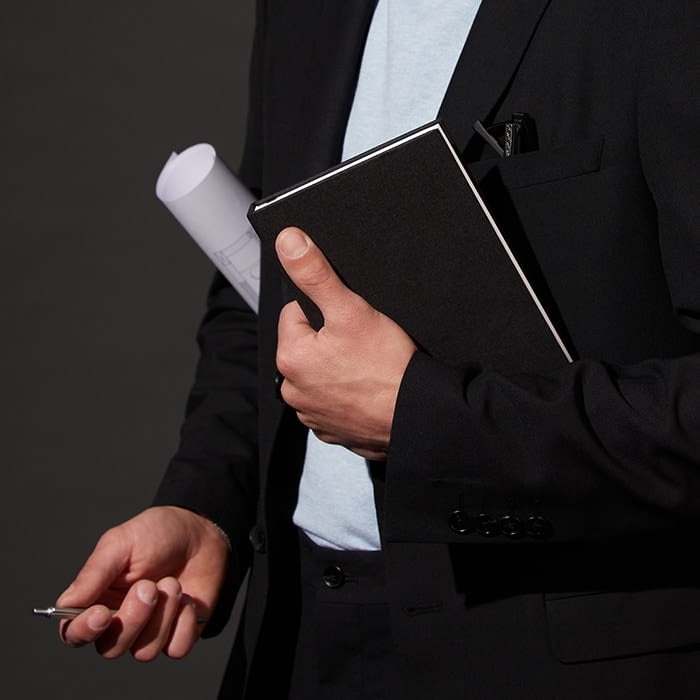 Person in a black suit holding a cloth hardcover notebook and rolled up blueprints in one hand and a key in the other