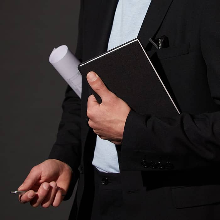 Person in a black suit holding a cloth hardcover notebook and rolled up blueprints in one hand and a key in the other