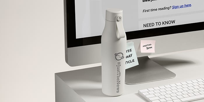 White custom water bottle with the 1440 logo and the hashtag Just The News on a desk next to a computer
