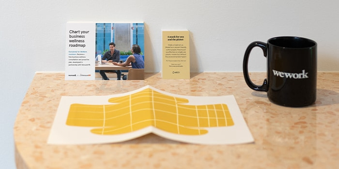 Yellow paper face mask on a desk with a postcard and a business card by MOO