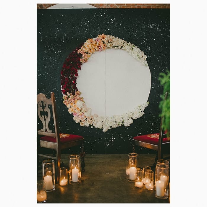 Black backdrop painted with white stars and with a big white rpund in the middle, covered in white, orange, and red flowers to form a crescent moon. Two vintage chairs and various candles are standing in front of the backdrop