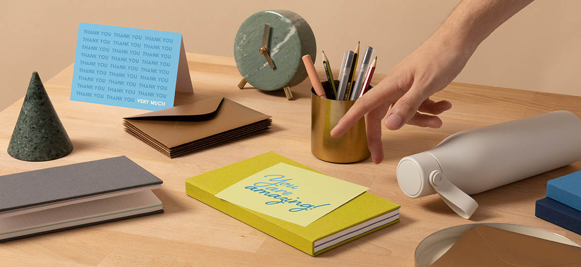 Hand pointing at You Are Amazing card on a desk next to a notebook, envelope, pencil pot and alarm clock
