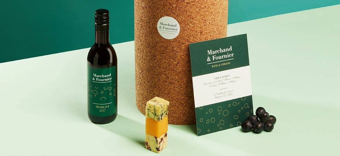 Mini bottle of wine with a green label, block of cheese, big cork pedestal with a round sticker, grape and big green postcard for a wine and cheese business