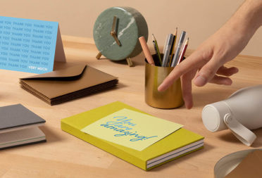 Hand pointing at You Are Amazing card on a desk next to a notebook, envelope, pencil pot and alarm clock