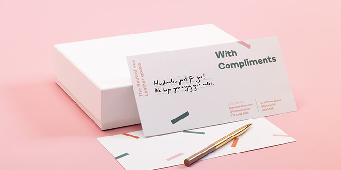 Pen and note card with handwritten message standing on a white box. The card says with compliments and makes use of white space with a minimalist design.