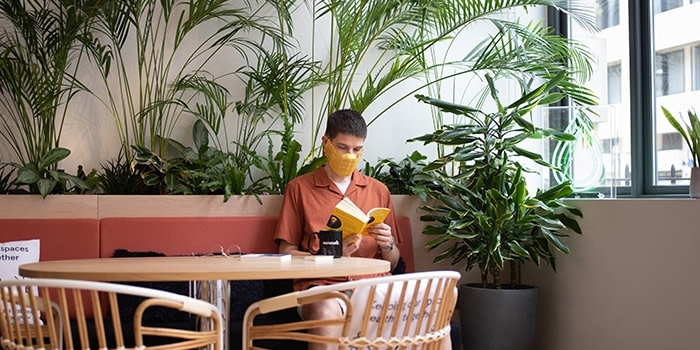 Person wearing a paper face mask and sitting at a table reading a book, with many plants in the background