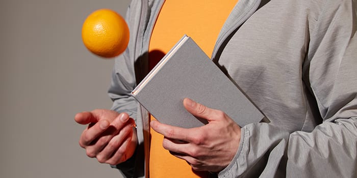 Person juggling with an orange and holding a light grey notebook