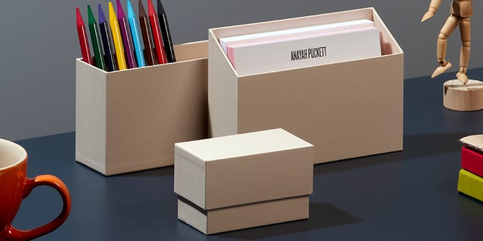 Beige display boxes in three sizes. One is closed, one is filled with postcards, and one is filled with pens
