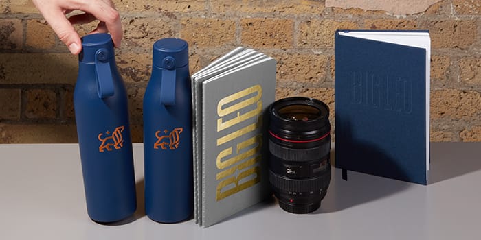 Branded Big Leo bottle and notebooks next to a camera lens