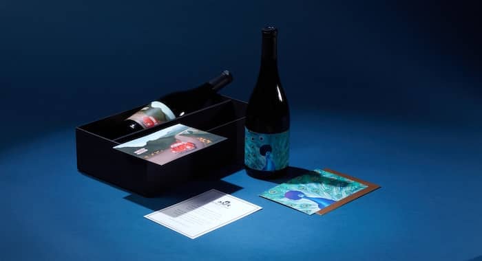 Open wine box with a bottle of McBride Sisters Reserve Collection wine with a red beetle car illustration on the label and a postcard with the same illustration. There is also another bottle with an afro peacock illustration, reflected on a premium postcard with a gold envelope.