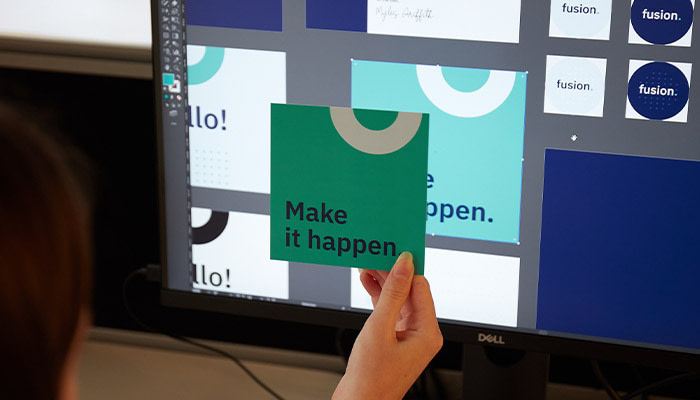 Holding up a square postcard with original design file on a computer in the background