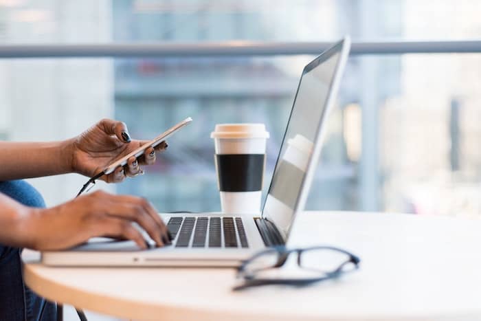 Hands typing on a laptop and looking at a phone. There is a takeaway cup and glasses on the desk.