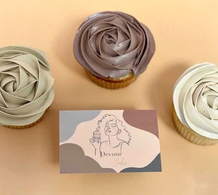3 cupcakes and a Devour cake shop business card with muted colors and a line drawing of a curly haired woman eating a cake