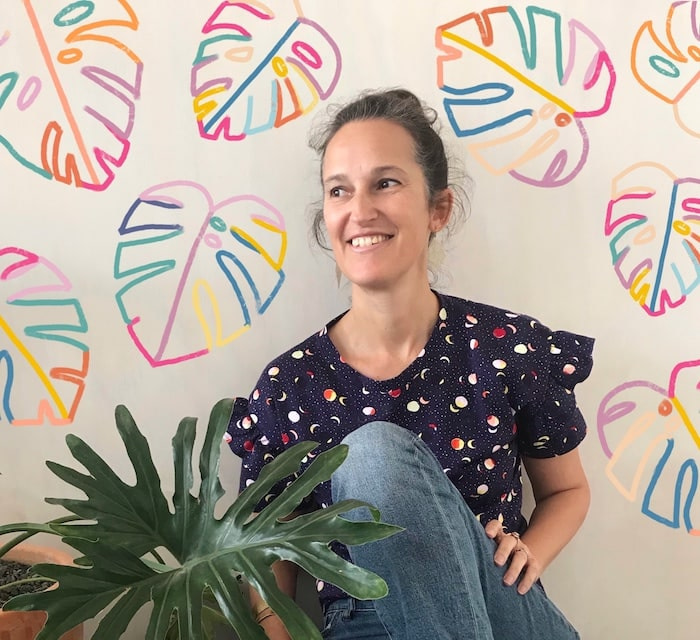 Portrait of surface designer Brook Gossen sitting next to a houseplant and smiling, with some of her colorful monstera pattern design in the background