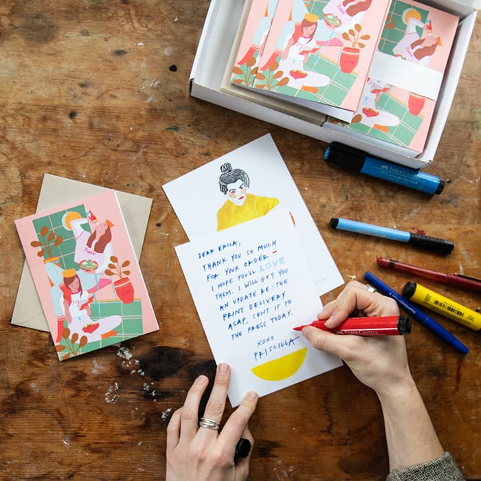 Priscilla Weidlein writing on a thank you card, surrounded by her holiday greeting card designs