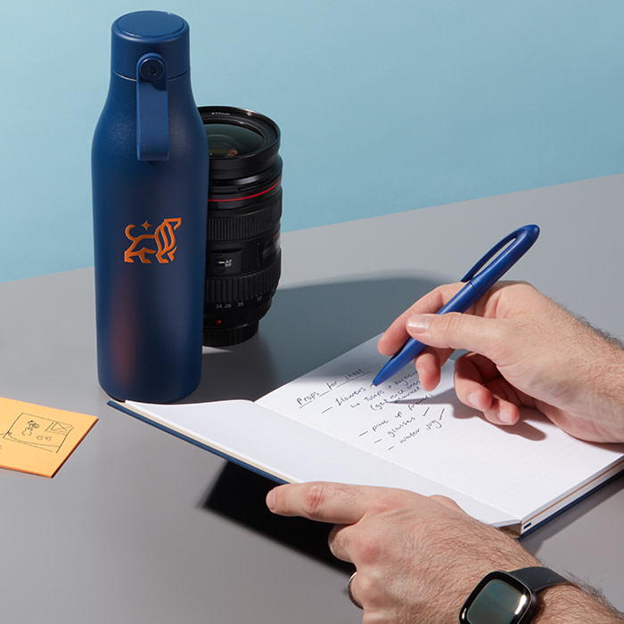 Hand writing into a blue Big leo notebook next to a Big Leo water bottle and a camera lens