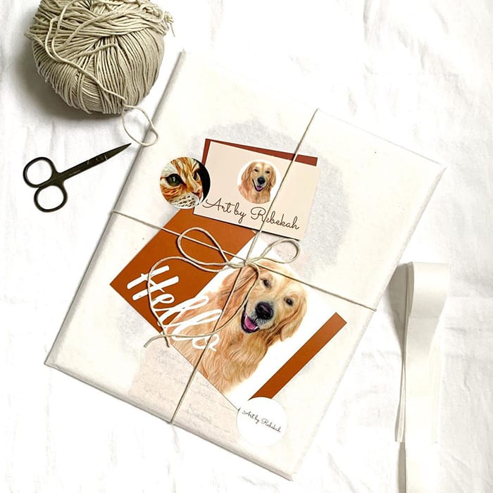 Ball of wool, scissors and packaging with a Hello dog postcard, a dog business card and a cat round sticker by pet portrait artist Rebekah Mushinski