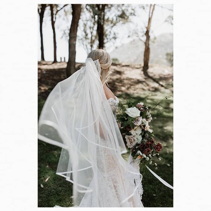 Bride with a big white veil seen from the back. She's standing in a clearing with trees in the background and she's holding a bouquet.