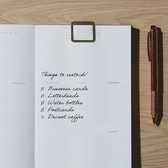 An open perpetual planner with a restock checklist written out. A brass planner clip is marking the page.